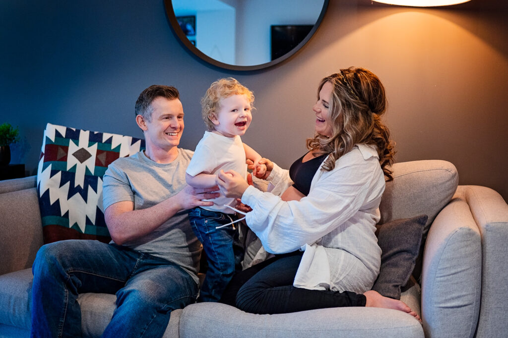 A joyful family lifestyle photoshoot in Glasgow captures a pregnant mother, father, and young child sitting on a cozy grey sofa in a warm, homey living room. The expectant mother, dressed comfortably, shares a laugh with her family as the child stands between them, adding a playful touch to the scene. A bouquet of colorful flowers adds warmth to the background, enhancing the relaxed and intimate atmosphere of the family session.