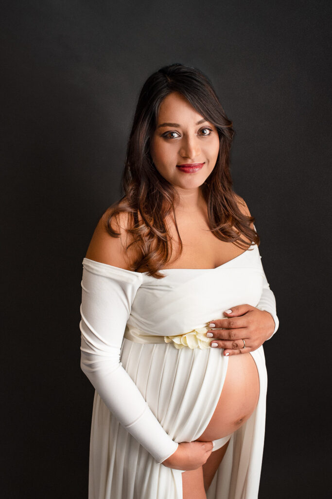 Pregnant woman in an off-the-shoulder white gown cradling her bare baby bump, standing against a dark background. She has long wavy brown hair and smiles softly, with one hand resting above her belly and the other below it during her maternity photography in Glasgow