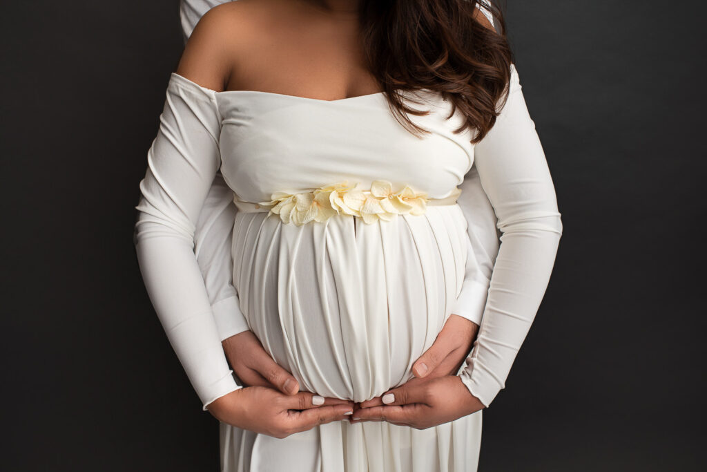 Pregnant woman in an off-the-shoulder white dress with floral detailing on the waist, standing in front of her partner. Both of their hands rest gently on her baby bump, creating a supportive and loving pose. The background is dark, drawing attention to the couple's hands and the belly.