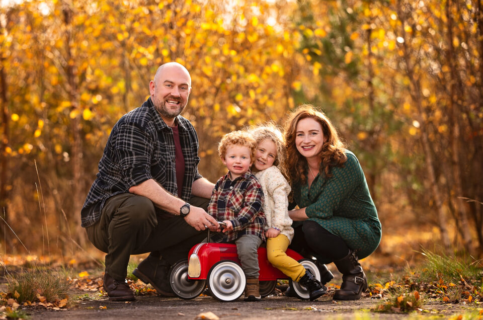 Happy family during an autumn outdoor photoshoot in Glasgow. Parents kneeling beside their two smiling young children sitting on a red vintage toy car, surrounded by golden autumn leaves and warm sunlight.