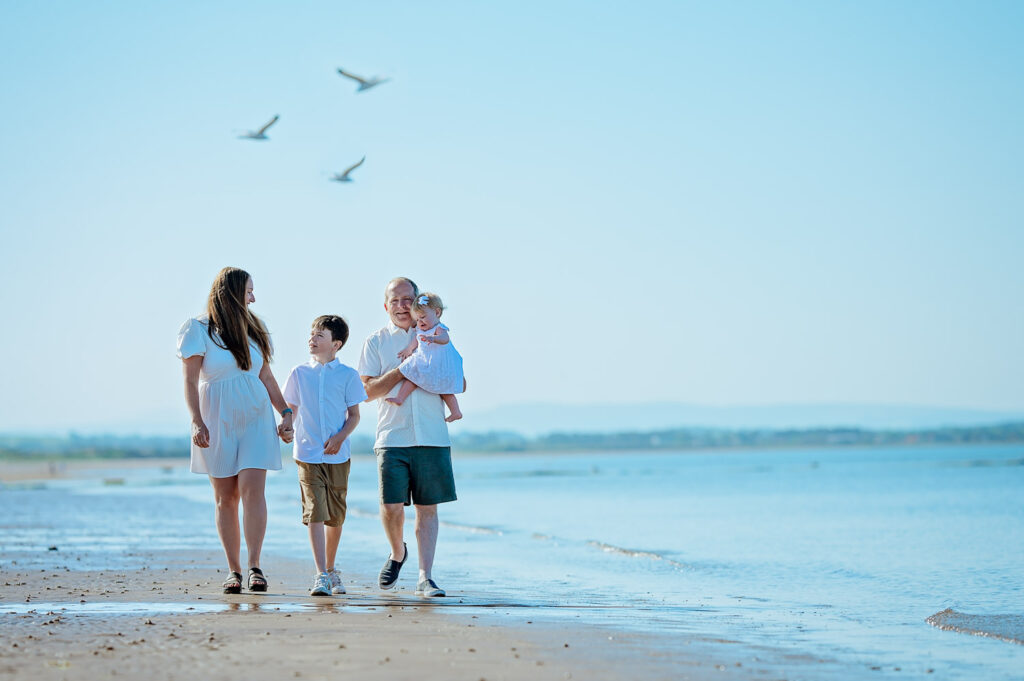Irvine Beach Family Photoshoot Family walking barefoot through shallow water during a sunny Irvine Beach family photoshoot in Scotland, captured by Ola Molik Photography